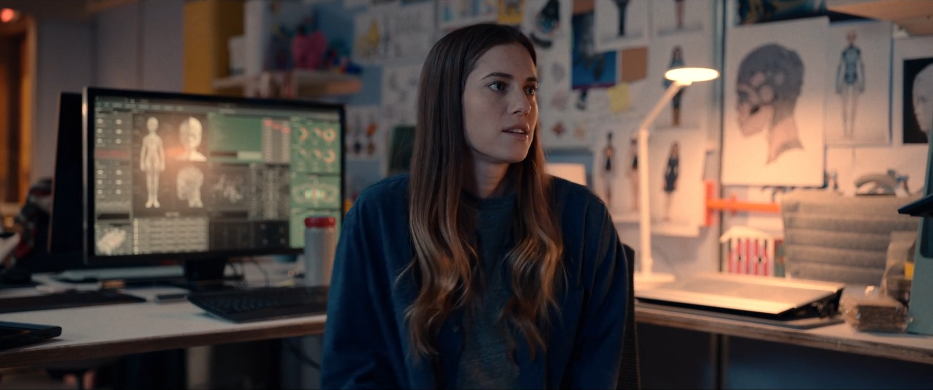 A woman with long brown hair and a blue jacket sitting at a desk in front of a computer with 3D medical images displayed. The background shows sketches and posters of human anatomy on the wall.