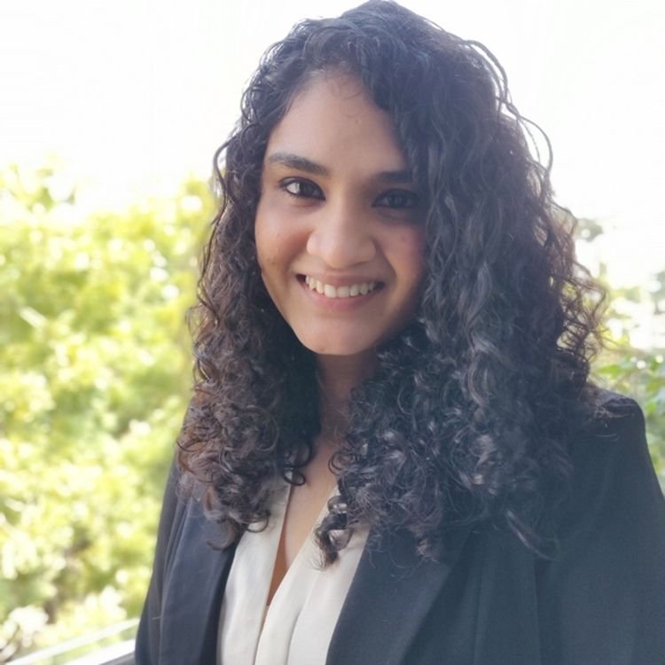 A young woman with curly dark hair smiling outdoors with green trees in the background.