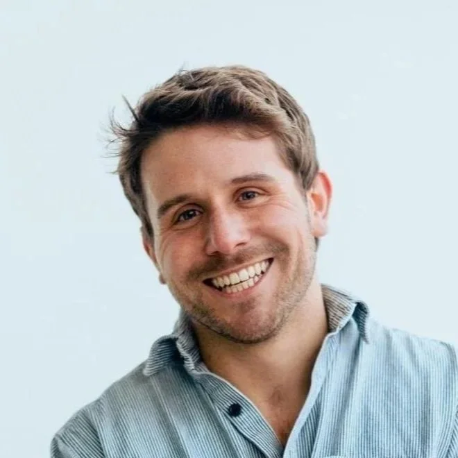 A man with short brown hair, a beard, and a big smile, wearing a light blue button-up shirt, standing against a plain light-colored background.