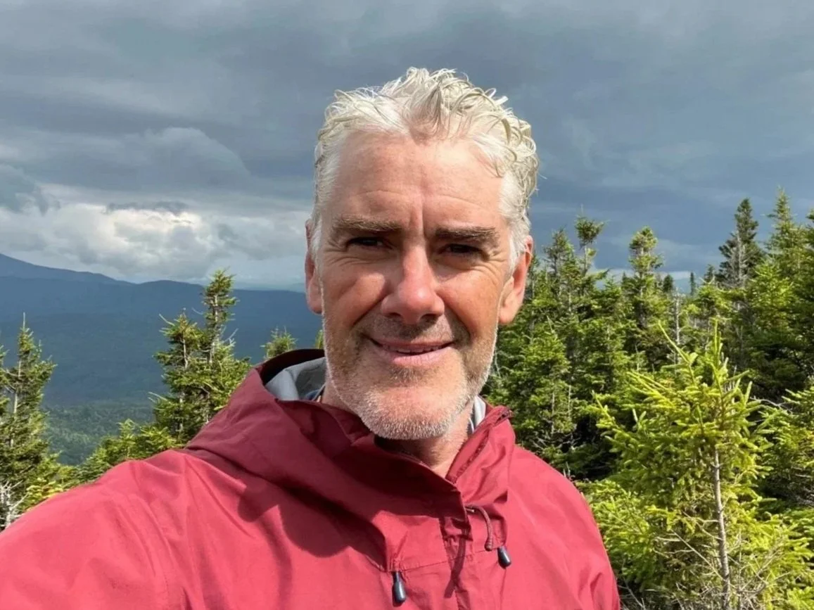 A man in a red jacket smiling outdoors with a mountain range and cloudy sky in the background.