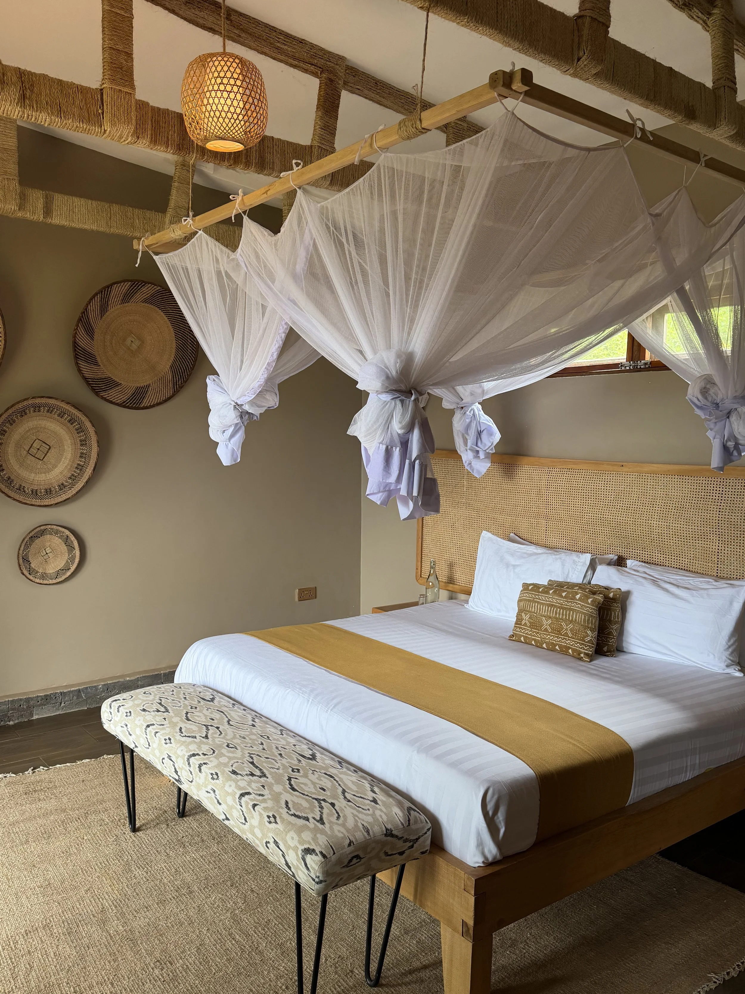 A neatly made bed with white sheets, brown and white pillows, and a yellow bed runner. A bench with patterned upholstery is at the foot of the bed. Hanging mosquito netting is above the bed, attached to wood and rope ceiling fixtures. Circular woven wall decorations are on the wall behind the bed, and a window with greenery outside is visible.