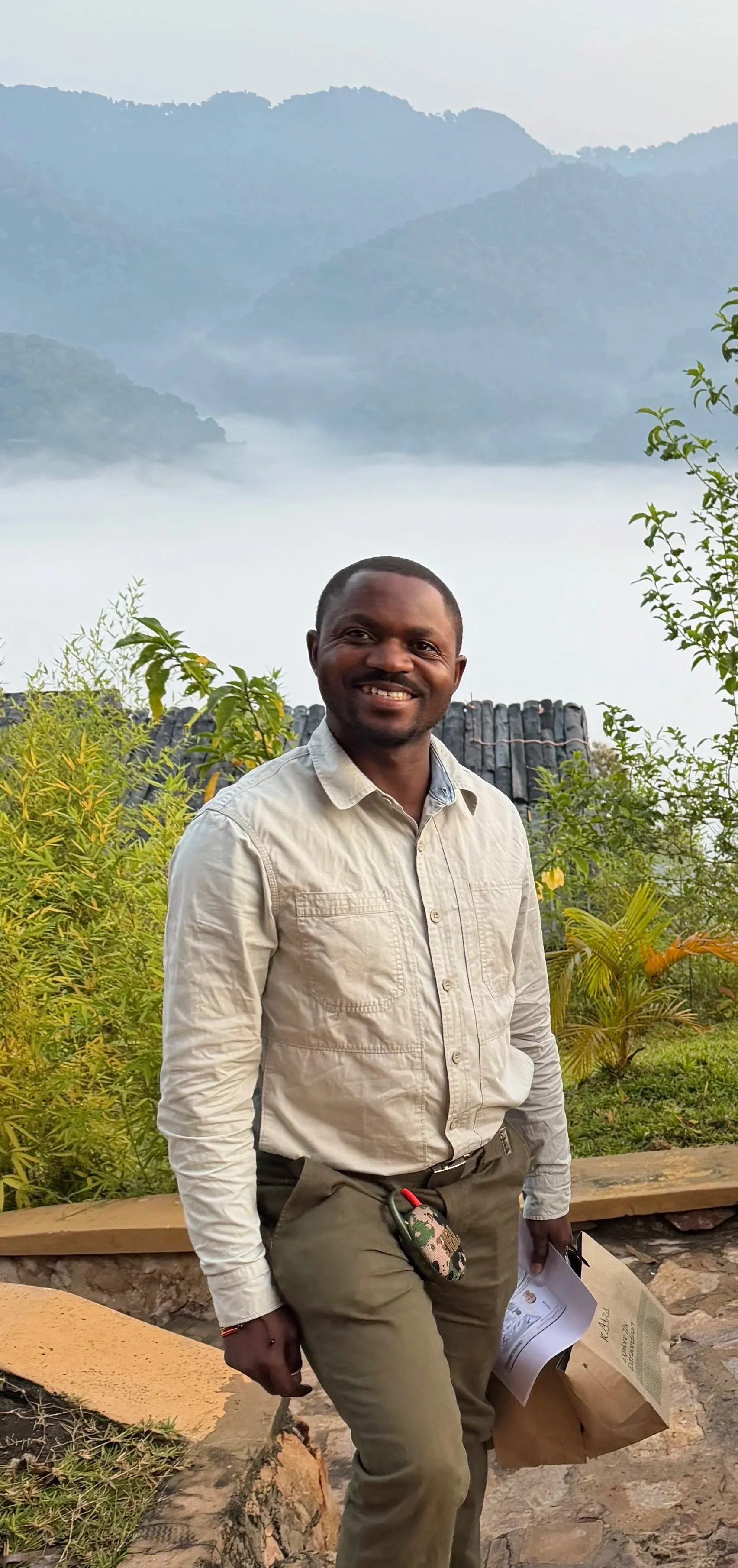 Baluku smiling wearing a beige shirt and khaki pants standing outdoors with greenery, mountains, and a misty lake in the background.