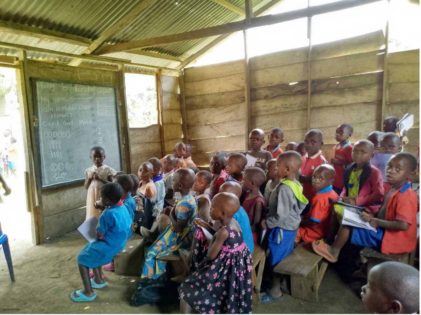 Children sitting in a classroom with a blackboard, inside a simple wooden structure with a corrugated metal roof.