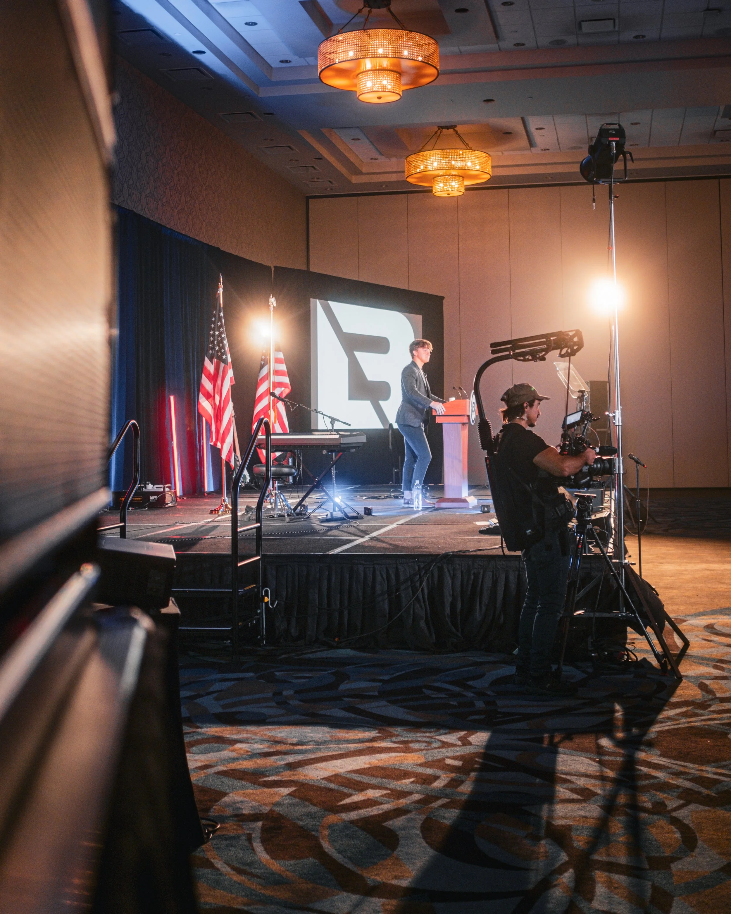 A person speaking at a podium on a stage with American flags behind them, during a conference or event. There are professional cameras filming the speaker, and stage lighting illuminating the scene.