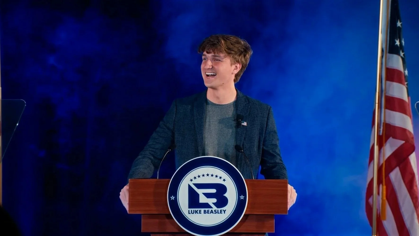 Young man with brown hair speaking at a podium with a logo reading 'LUKE BEASLEY' and a stylized 'LB' symbol. The background is blue, and an American flag is visible on the right.