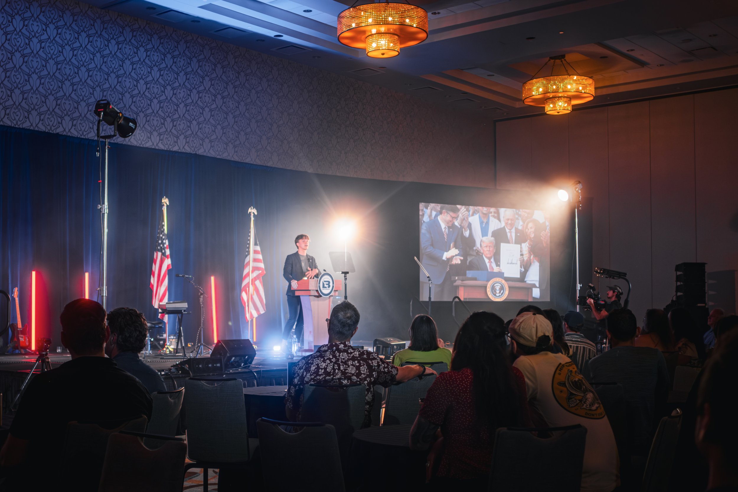 A woman speaking at a podium in front of an audience at a formal event or conference. There are two American flags behind her, and a large screen projecting her image. The setting is a dimly lit ballroom with chandeliers on the ceiling.