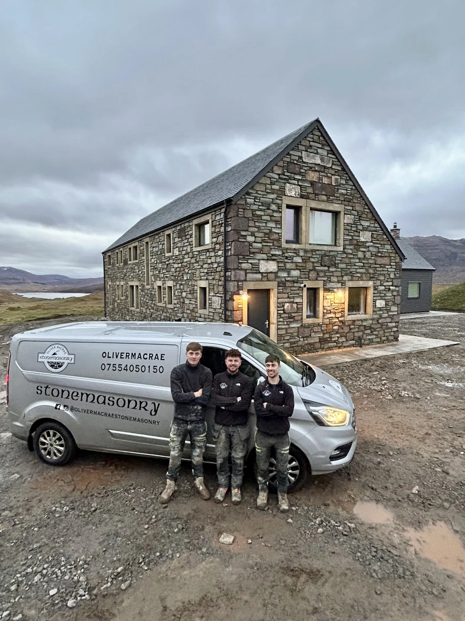 Three men standing in front of a garage door beside a gray van with the text "stonemasonry" and "Oliver Macrae" on it, in front of a stone house in a rural area with mountains and water in the background.
