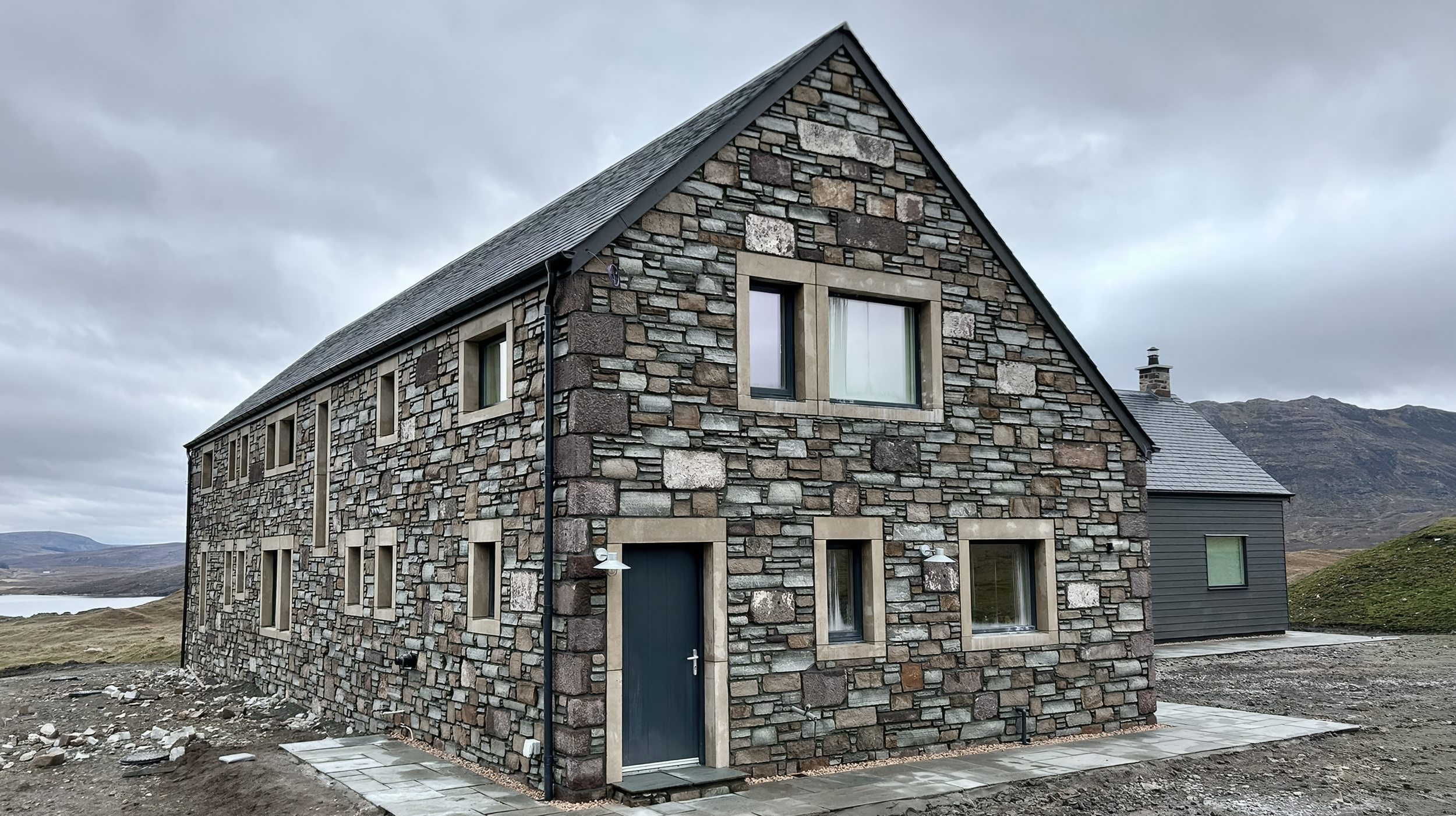 A modern stone house with multiple windows, a dark door, and a sloped roof, set in a mountainous landscape with cloudy skies.