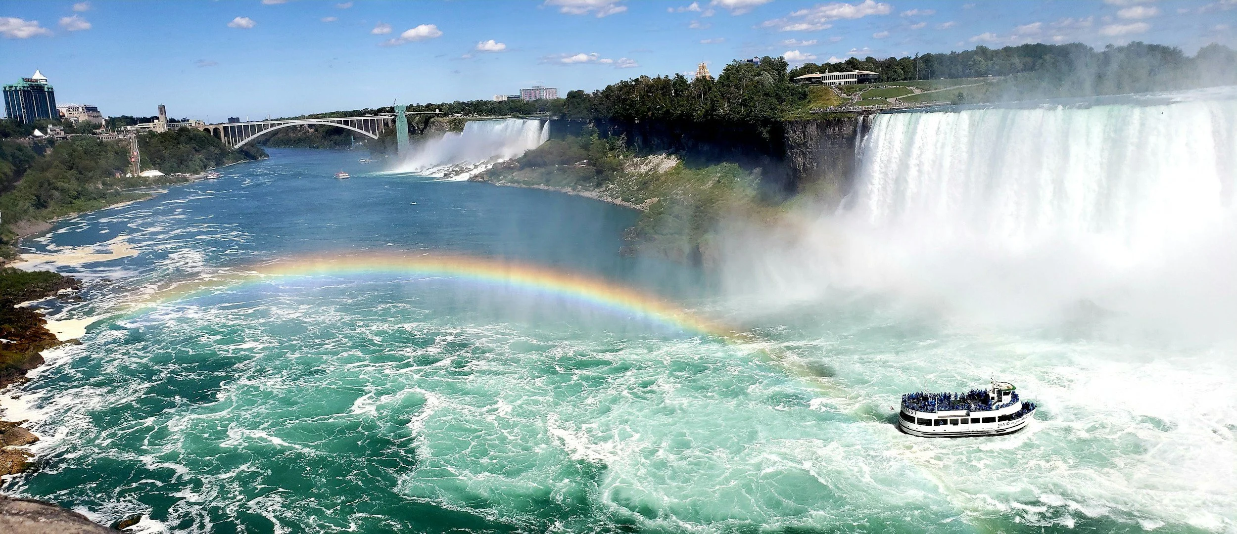 View of Niagara Falls with a rainbow in the mist, a boat in the river, and cityscape with buildings and a bridge in the background.