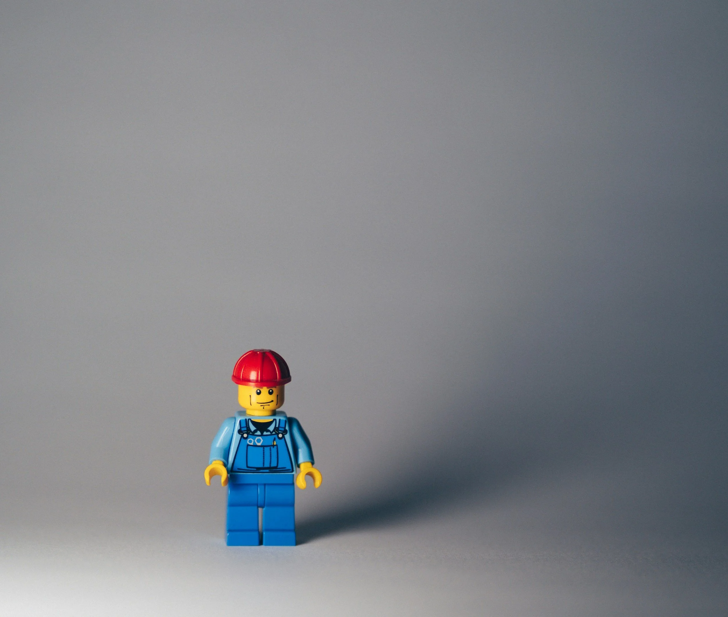 A LEGO minifigure dressed as a construction worker with a red helmet, blue overalls, and a neutral expression, standing on a gray surface with a gray background.