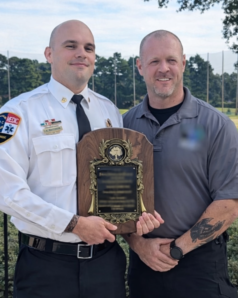 Two men standing outside, holding a wooden plaque with a gold emblem and black text. The man on the left is dressed in a white uniform with a badge and medals, and the man on the right is wearing a gray polo shirt with a tattoo on his arm.
