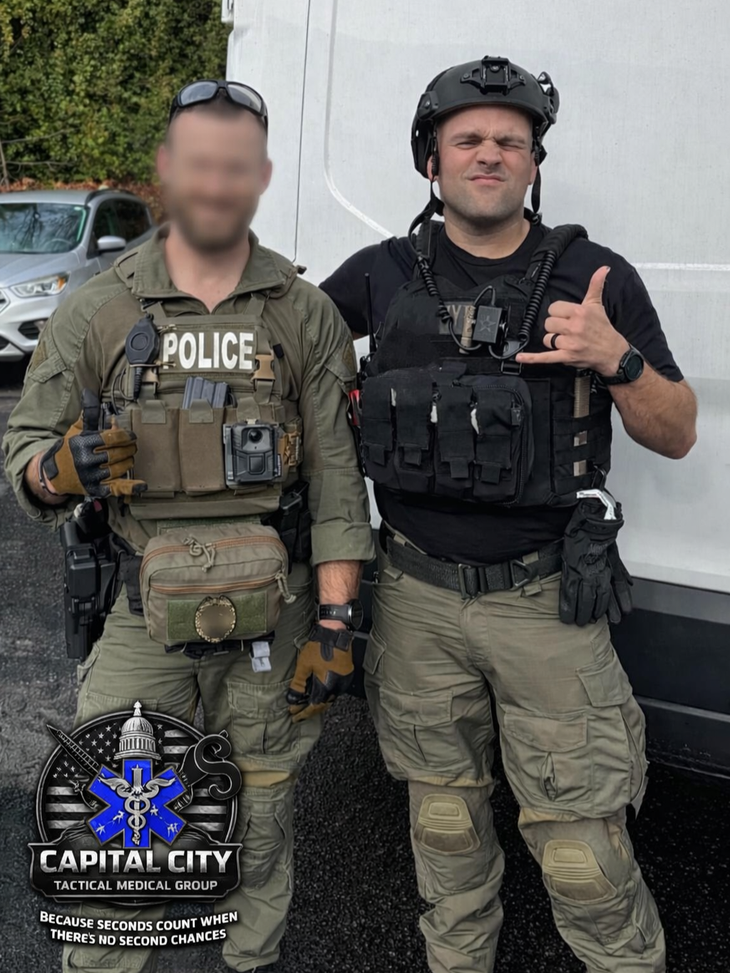 Two tactical medical group members in tactical gear, one dressed as police, standing outside in front of a white wall. One male is making a shaka sign with his hand, wearing a helmet, tactical vest, and gloves, while the other is standing with a blurred face, wearing sunglasses on his head and a tactical vest with a badge saying 'POLICE'.