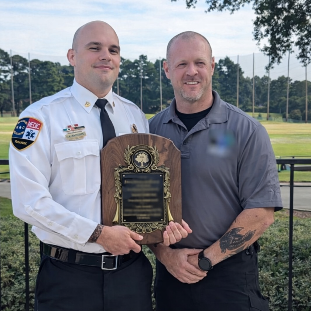 Two men standing outdoors, one in a fire department uniform holding a plaque, the other in a gray shirt with tattoos on his arm, on a grassy area with trees and a baseball field in the background.