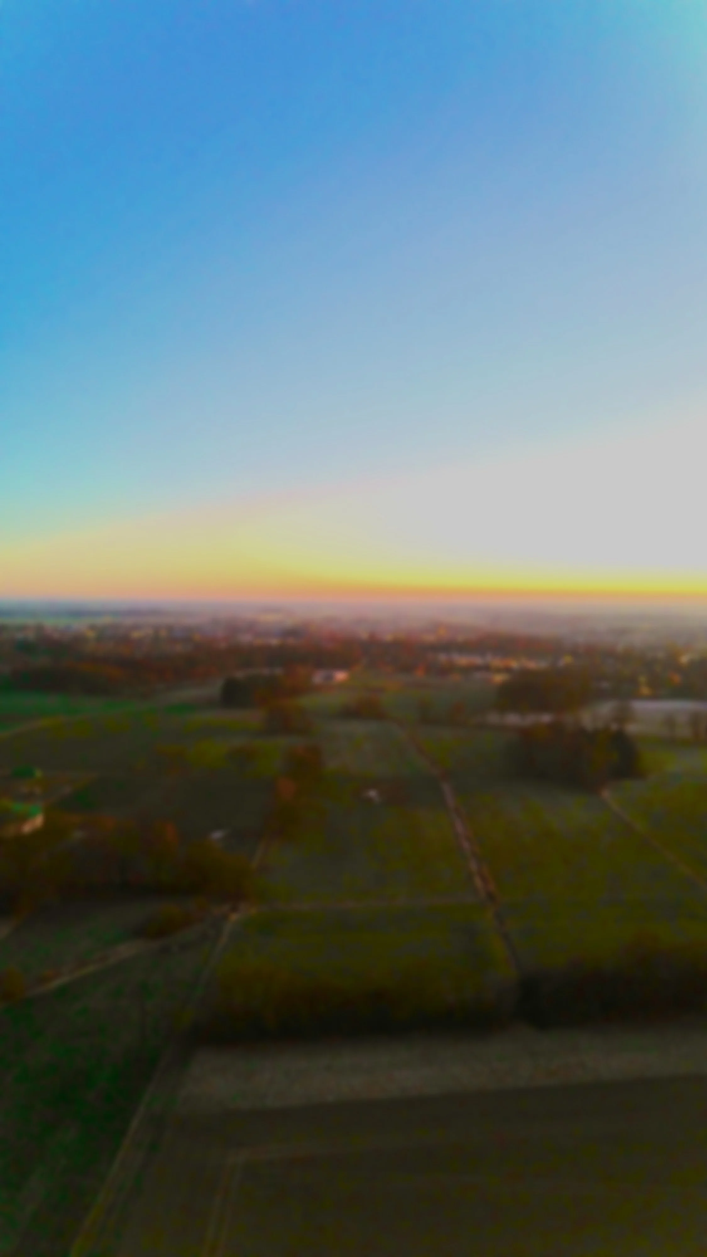 Landschaftsaufnahme mit Feldern, Bäumen und einem Himmel bei Sonnenuntergang oder Sonnenaufgang.