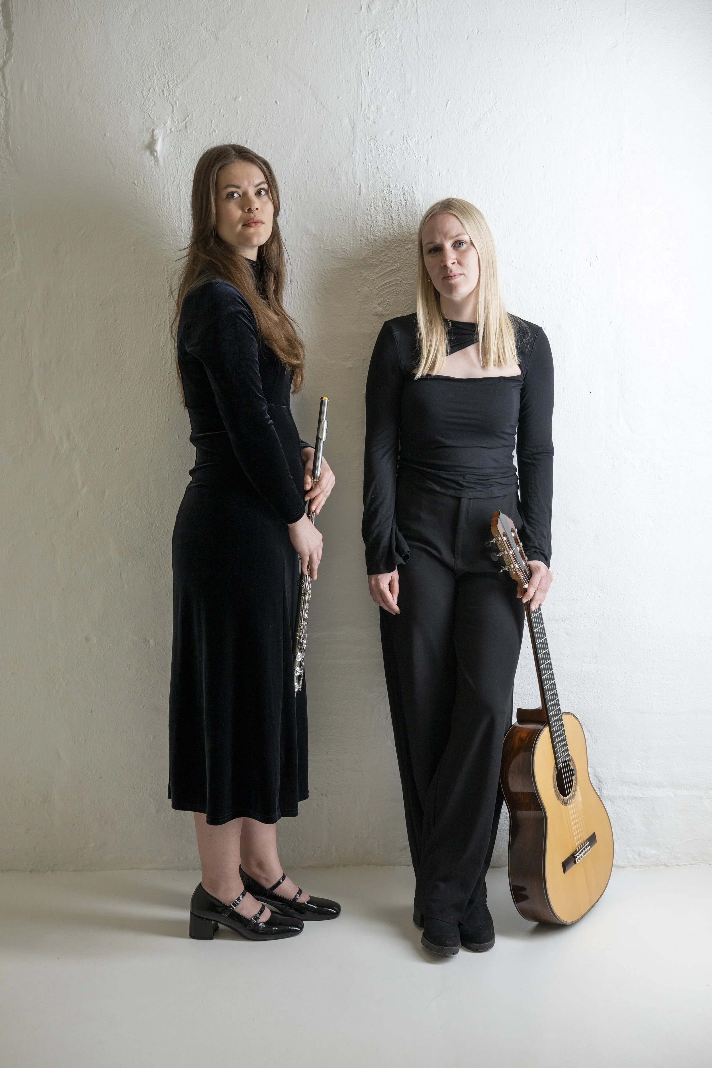 Två kvinnor står bredvid varandra mot en vit vägg, den ena håller en flöjt och den andra en akustisk gitarr, båda klädda i svarta kläder.