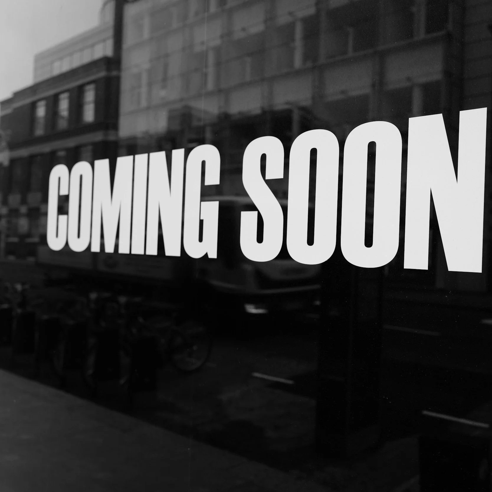 Black and white storefront window with large text saying 'COMING SOON'; reflections of buildings and bicycles visible in the glass.