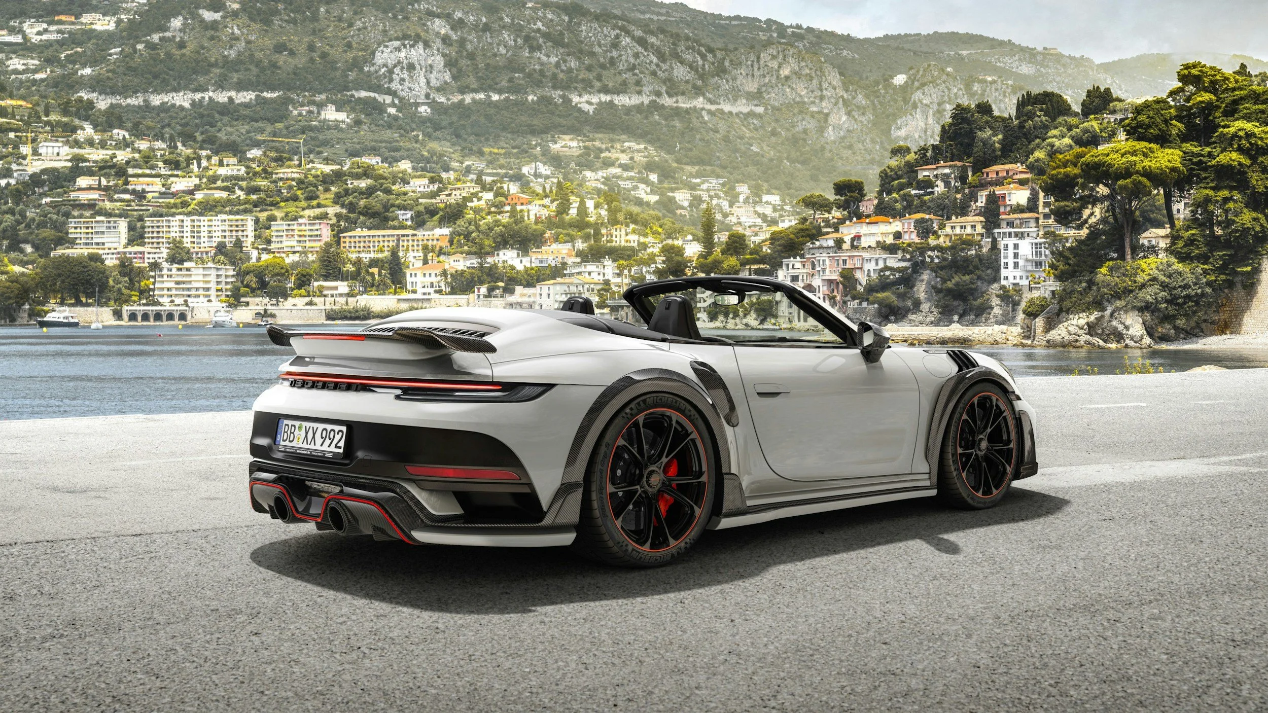 A silver sports car with black and red accents parked by a waterfront with hills and town buildings in the background.