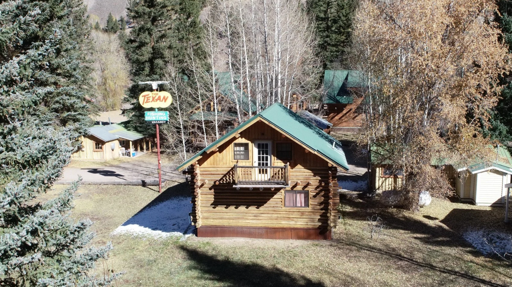 A wooden cabin with a green metal roof, a small front porch, and a window, situated in a wooded area with trees and patches of snow on the ground. There is a sign for The Texan motel that mentions fishing, hunting, and vacancies.