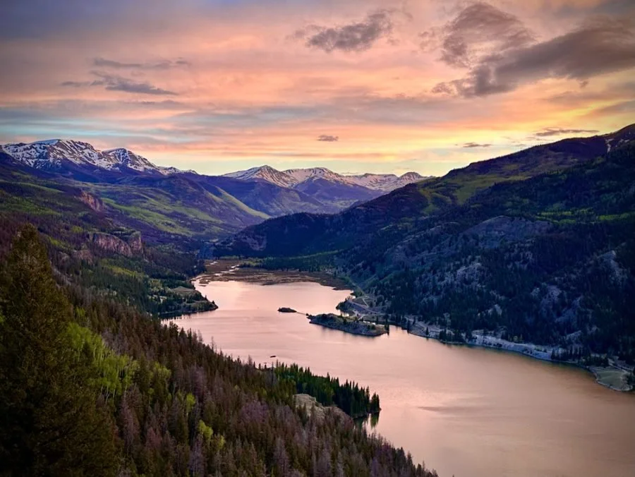 Scenic view of a winding river through green forested mountains during sunset with snow-capped peaks in the background.