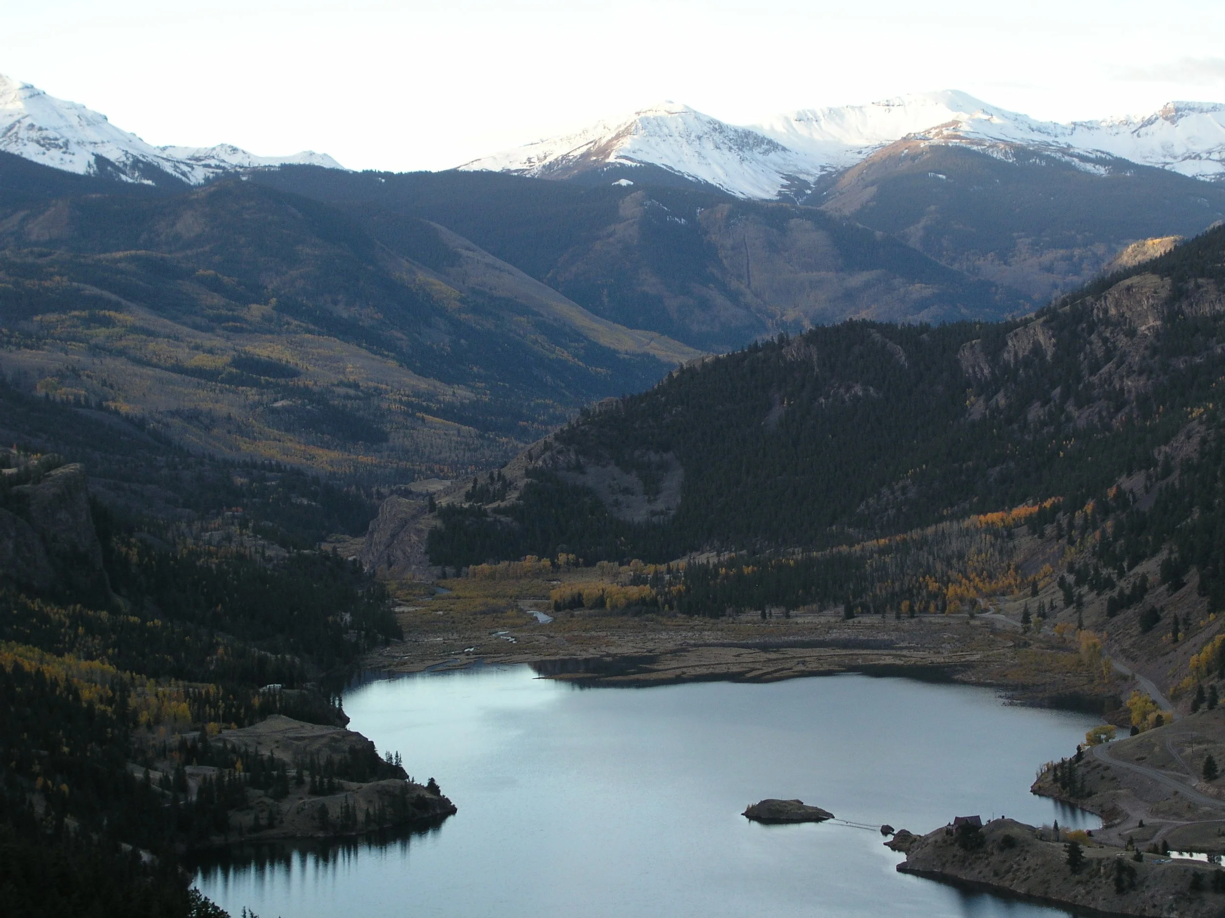 A scenic mountain landscape with snow-capped peaks, forested slopes, and a calm lake in the foreground.