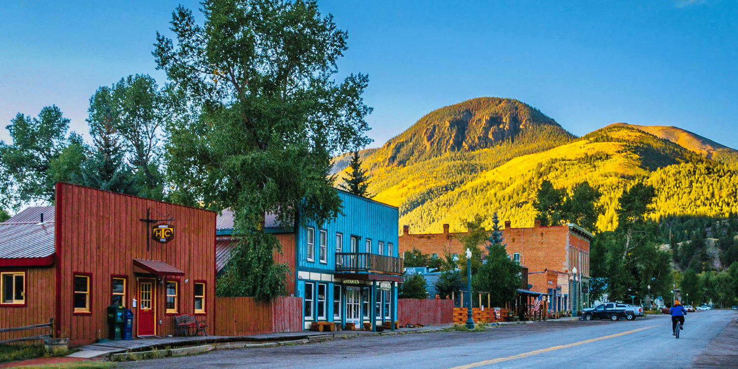 A small town street with colorful buildings, trees, and mountains in the background during sunset.