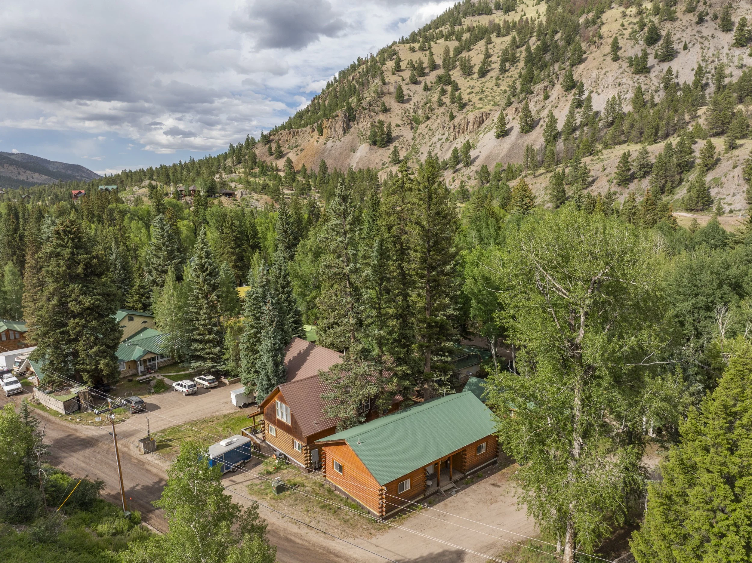 Aerial view of a small mountain town with wooden houses, some with green and brown roofs, nestled among tall trees. The background features a forested hillside with sparse trees and a cloudy sky.