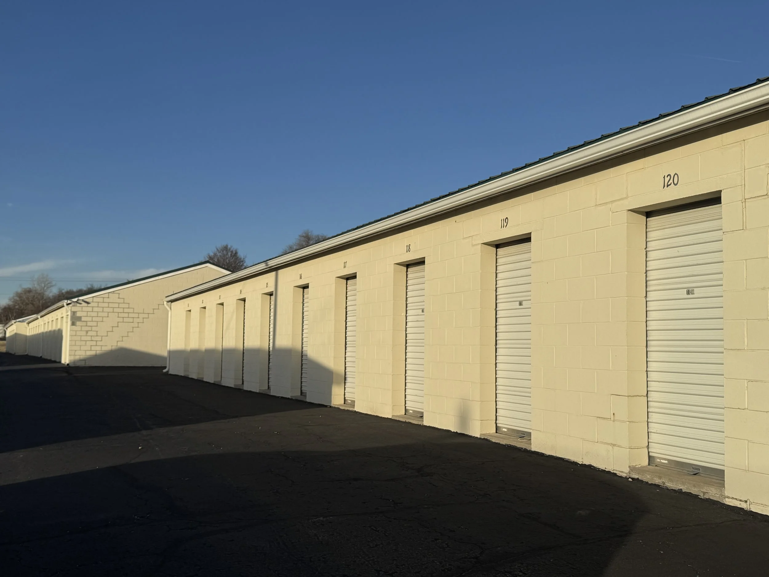 Row of beige storage units with closed roll-up doors, each numbered, under a clear blue sky.