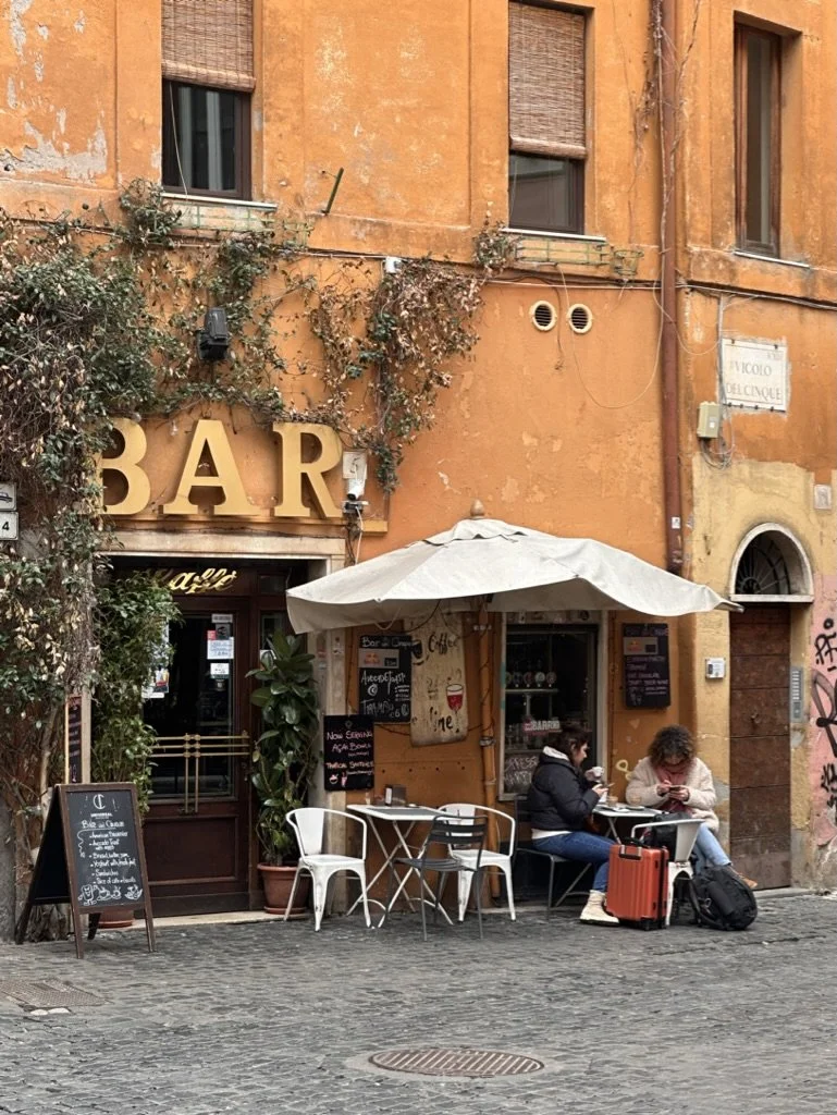 Two ladies traveling sitting at an outdoor table with an umbrella at a quaint little bar.