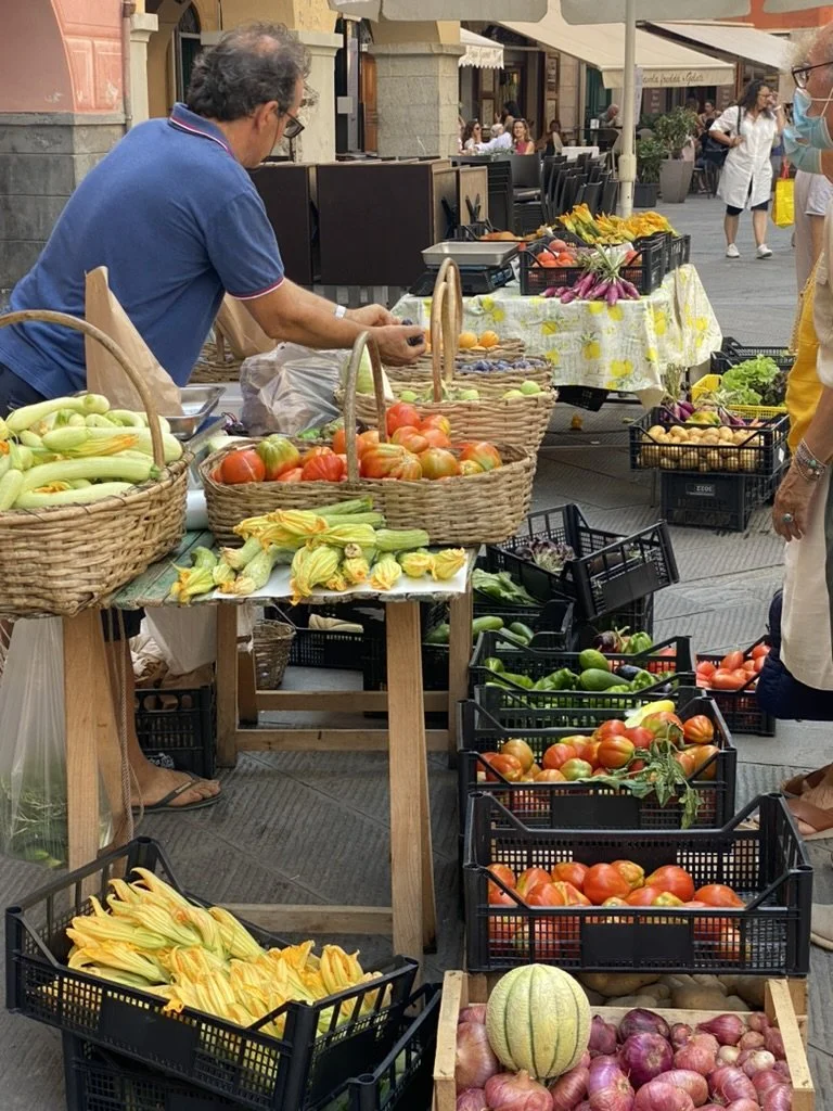 Man at his fruit and vegetable stand in Italy, as travelers stroll by.