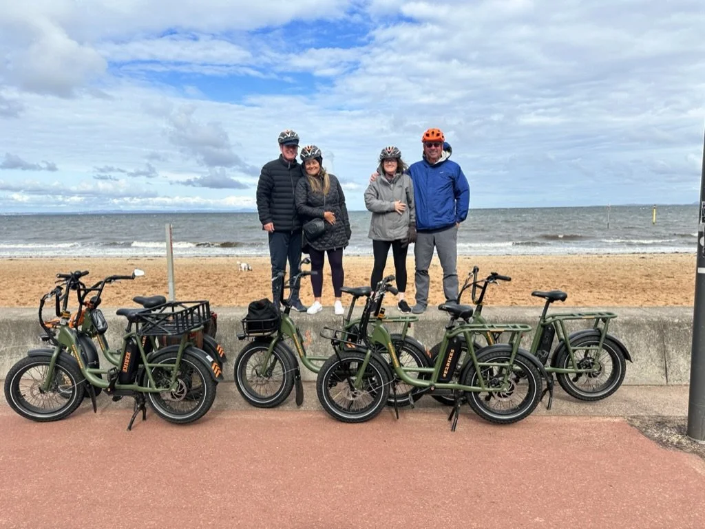 Two couples standing on a beach boardwalk, with the ocean behind them, and four e-bikes in front of them.