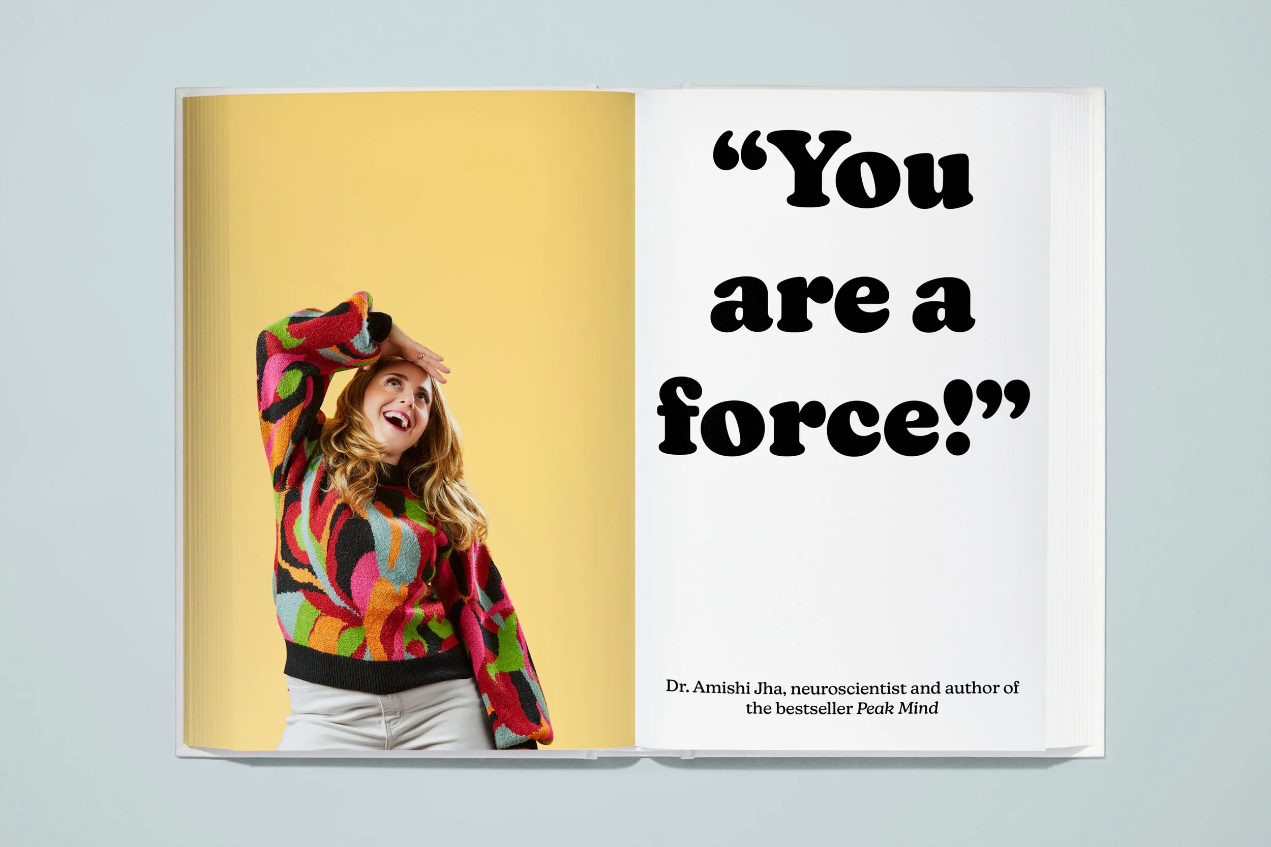 Open book showing a woman in a colorful patterned sweater with a yellow background on the left page and a quote in bold black letters on the right page that reads 'You are a force!' along with a smaller caption crediting Dr. Amishi Jha.
