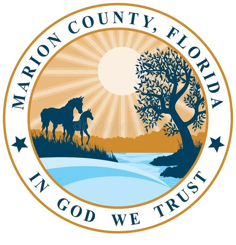 Seal of Marion County, Florida, featuring two horses near a river, a large tree, the sun shining, with the words "Marion County, Florida" and "In God We Trust" around the border.