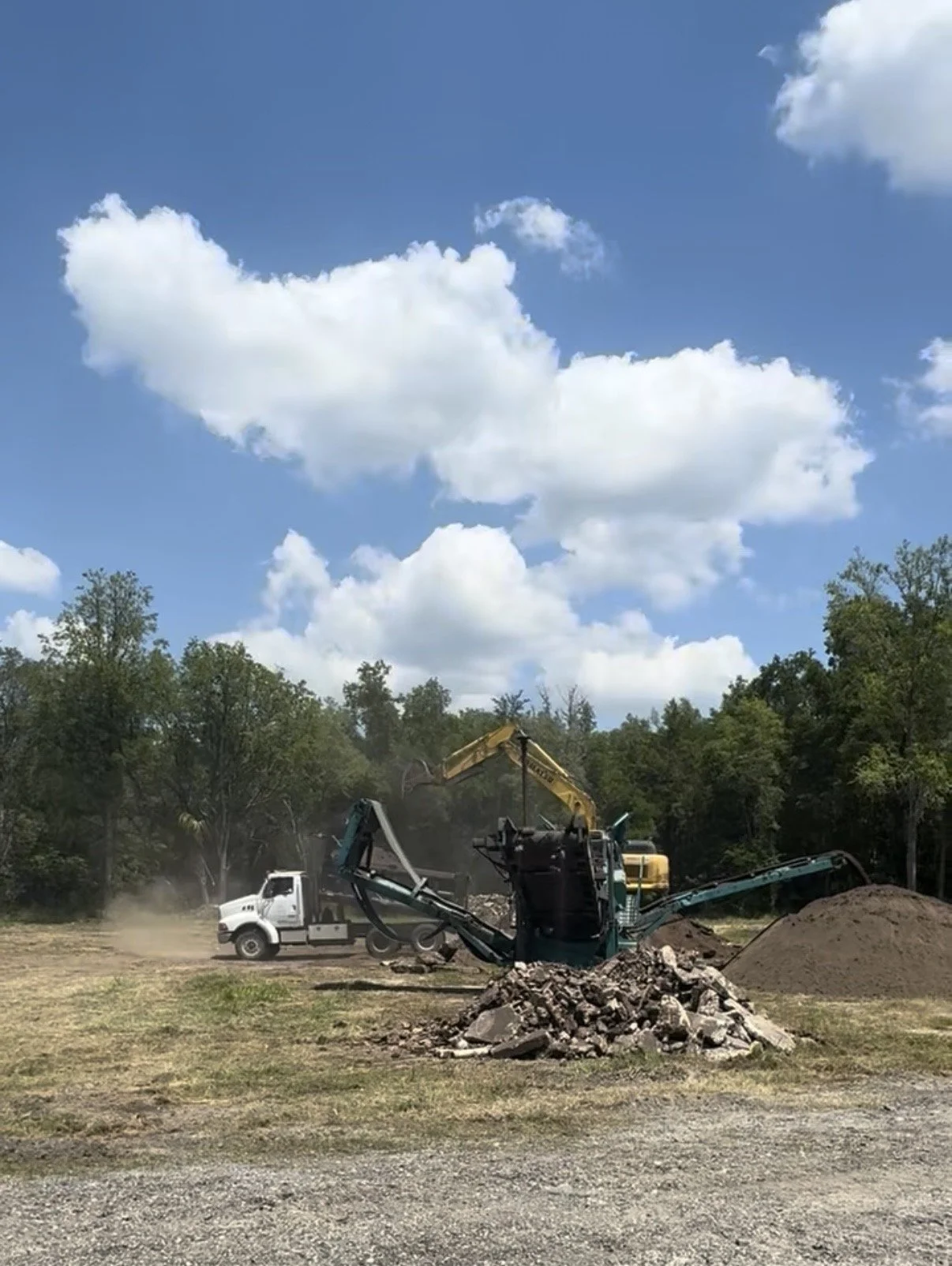 Construction site with excavator, dump truck, piles of dirt, and stones under blue sky with clouds.