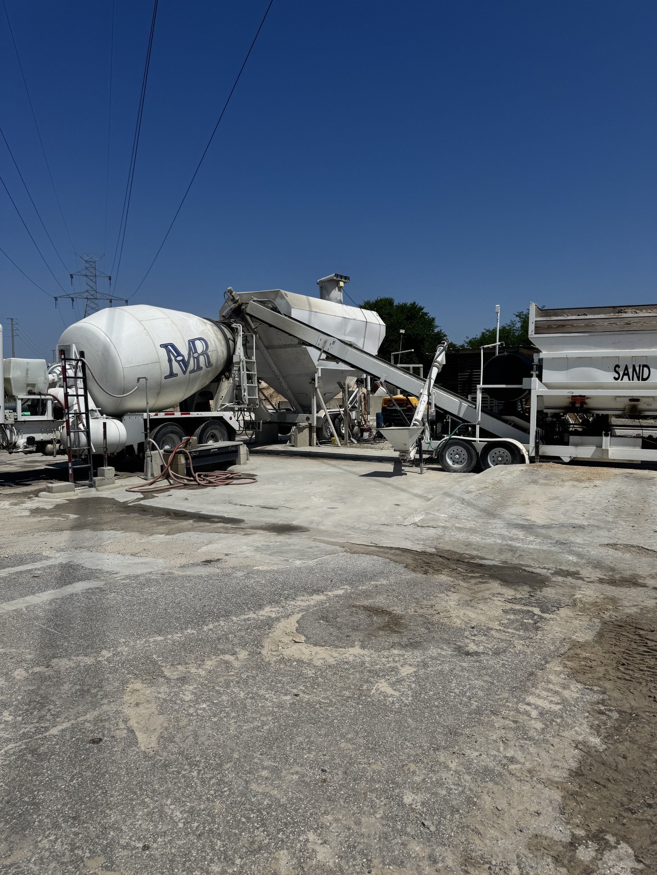 Construction site with cement mixers labeled 'MR' and 'SAND', concrete and equipment under a clear blue sky.