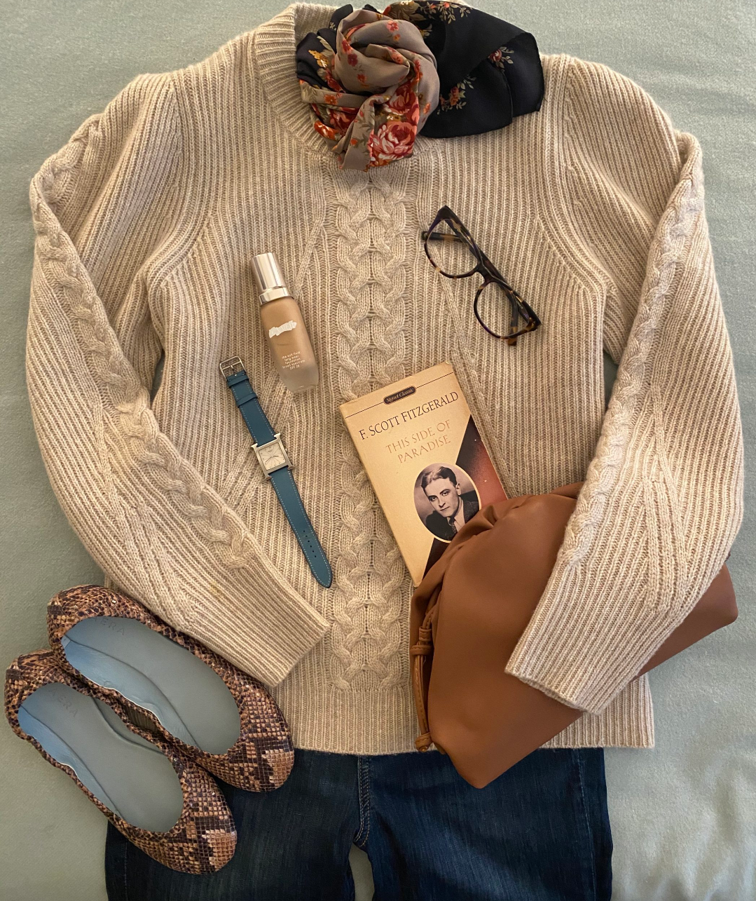Flat lay of a cozy outfit including a cream cable-knit sweater with jeans, a pair of patterned flats, a blue watch, a bottle of lotion, a brown bag, a pair of glasses, a book titled 'This Side of Paradise' by F. Scott Fitzgerald, a black and floral scarf, and a beige knit scarf.