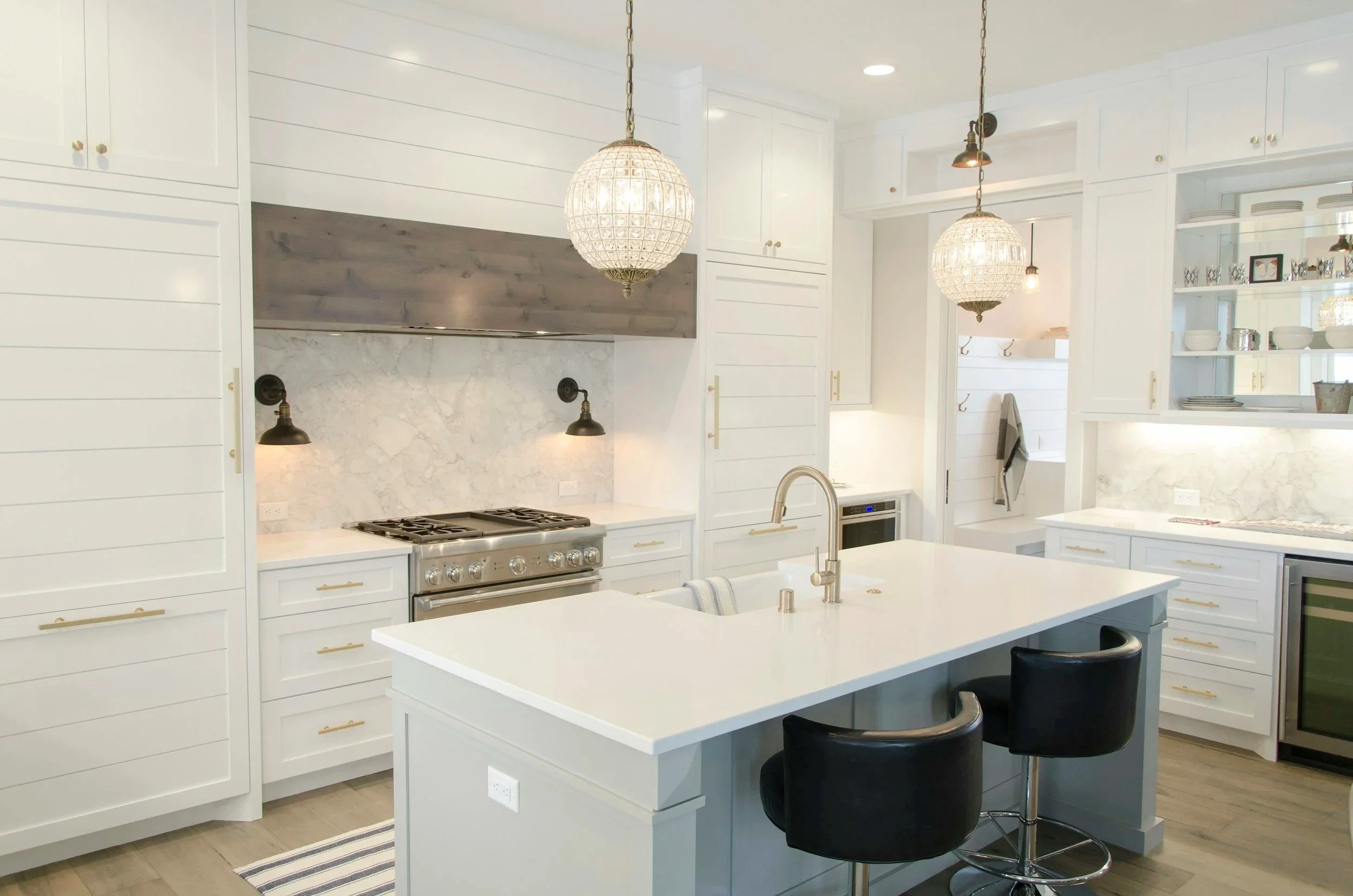 Modern white kitchen with island, two black chairs, pendant lighting, and open shelving.