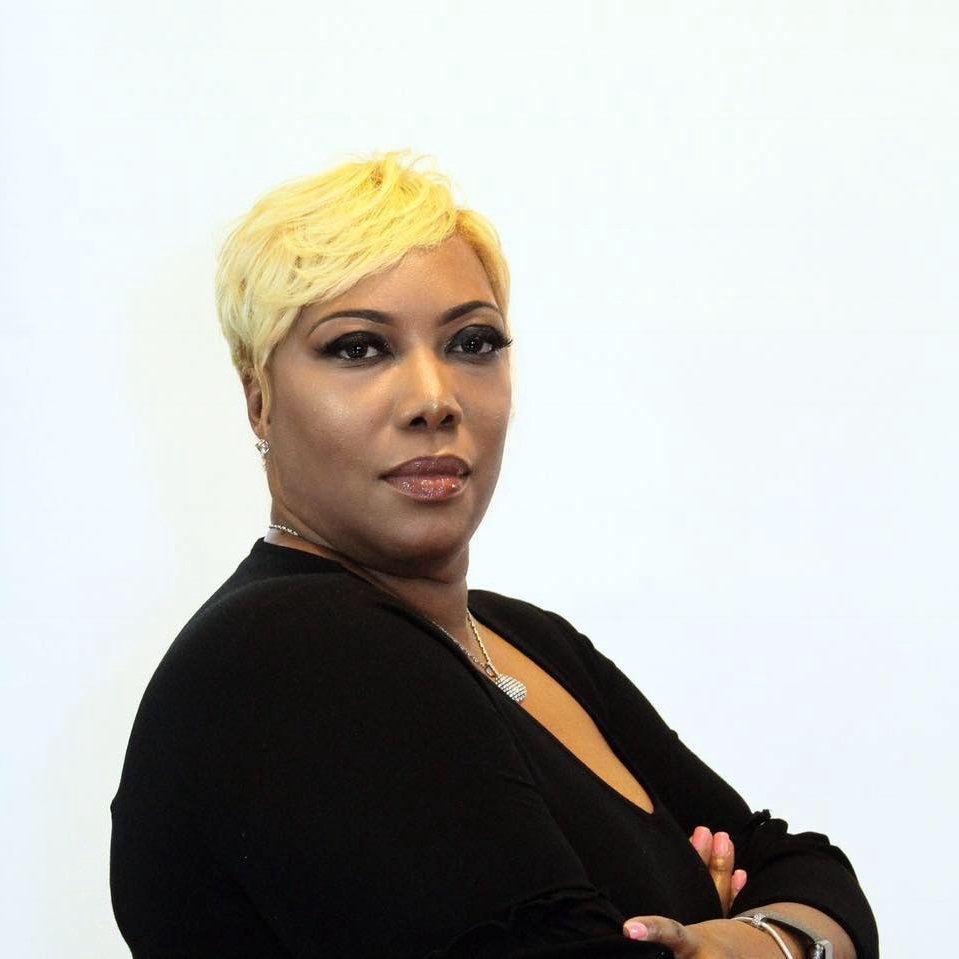 Portrait of a woman with short, blonde hair, wearing a black top and jewelry, standing against a plain white background.