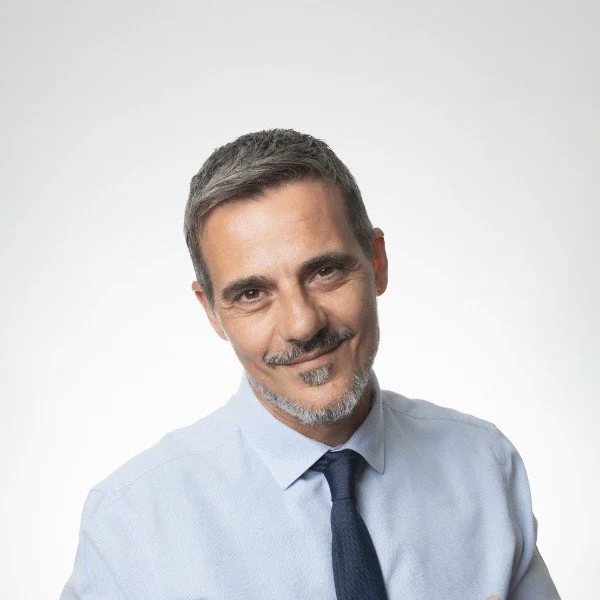 Close-up of a middle-aged man with short, gray hair and a beard, wearing a light blue dress shirt and a dark tie, smiling slightly, against a plain white background.