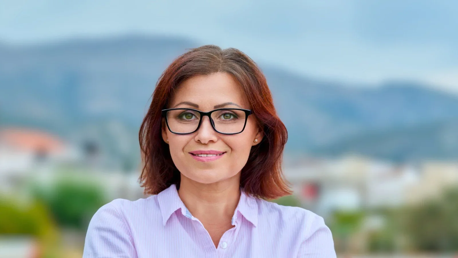 Close-up of a smiling woman with auburn hair and glasses outdoors, with a blurred background of mountains and buildings.
