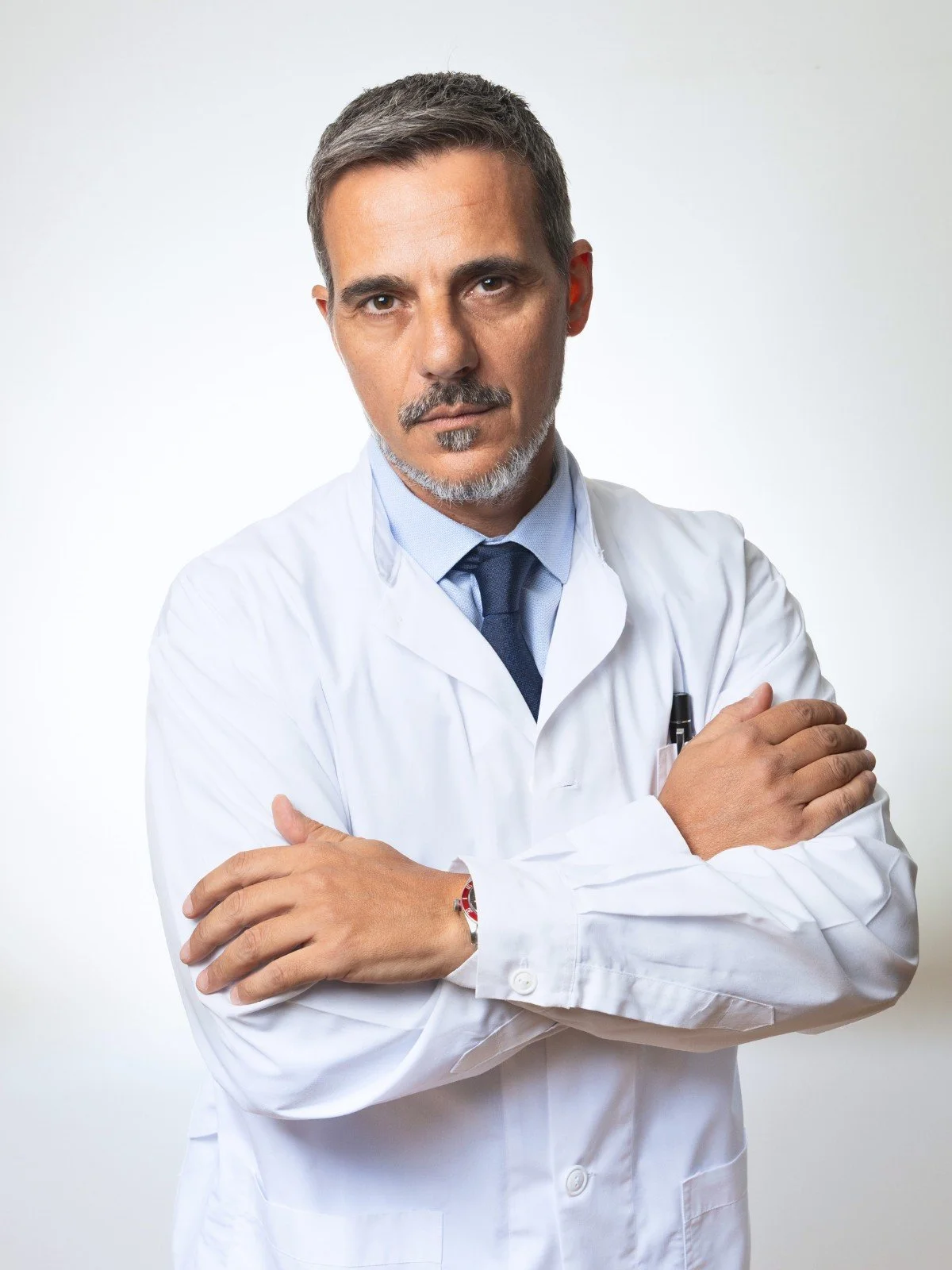 A middle-aged male doctor with short dark hair and a mustache, wearing a white lab coat, a light blue shirt, and a dark tie, standing with arms crossed against a plain white background.