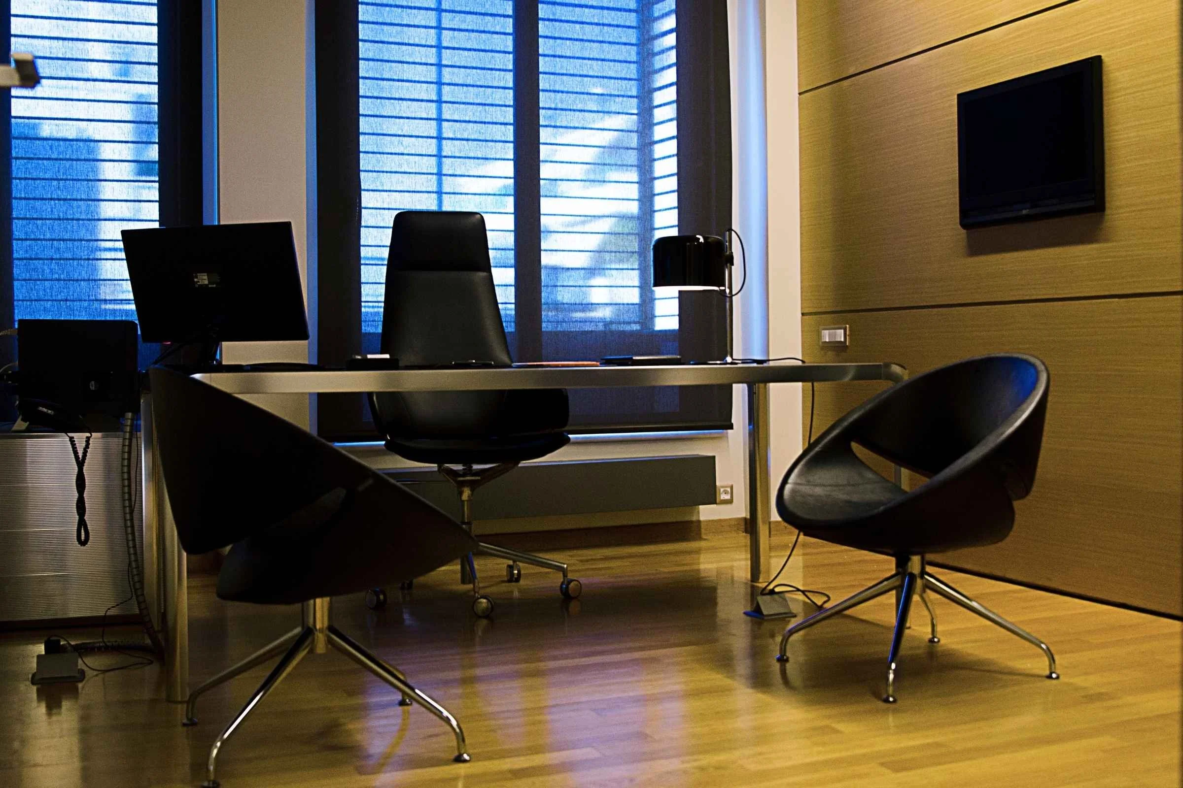 Modern office with a desk, three black chairs, a computer, a black coffee machine, and a large window with blue blinds.