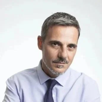A middle-aged man with short gray hair and a beard, wearing a light blue dress shirt and a dark blue tie, posing against a plain background.