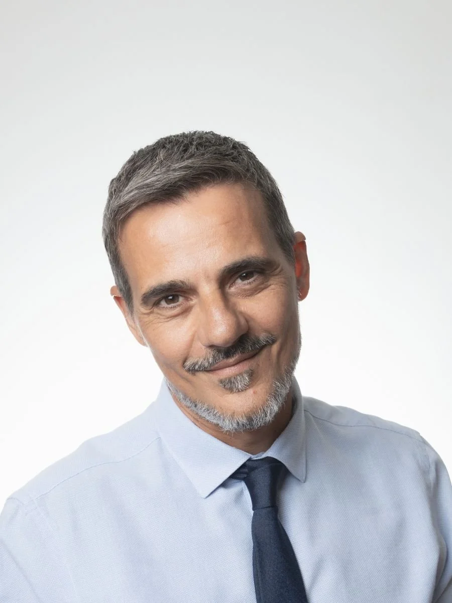 Smiling man with short gray hair, a beard, dressed in a light blue shirt and dark tie, posing against a plain white background.