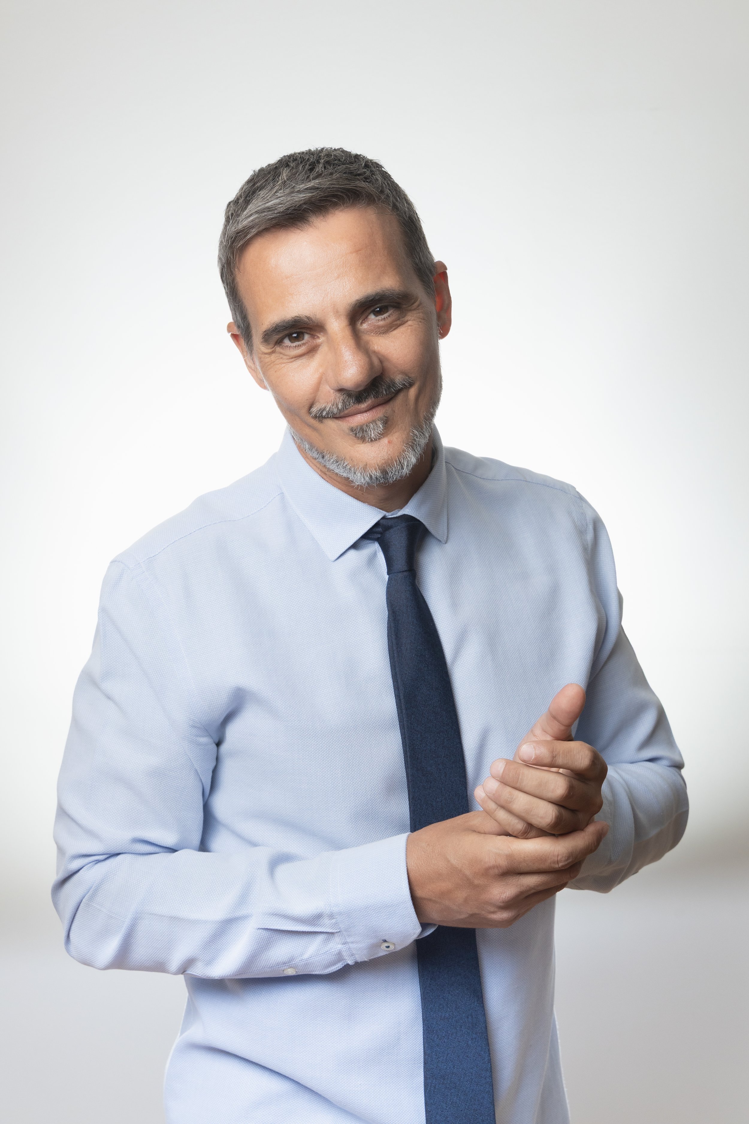 A middle-aged man with salt-and-pepper hair and a beard, wearing a light blue dress shirt and navy tie, smiling and clasping his hands together while looking at the camera against a plain white background.