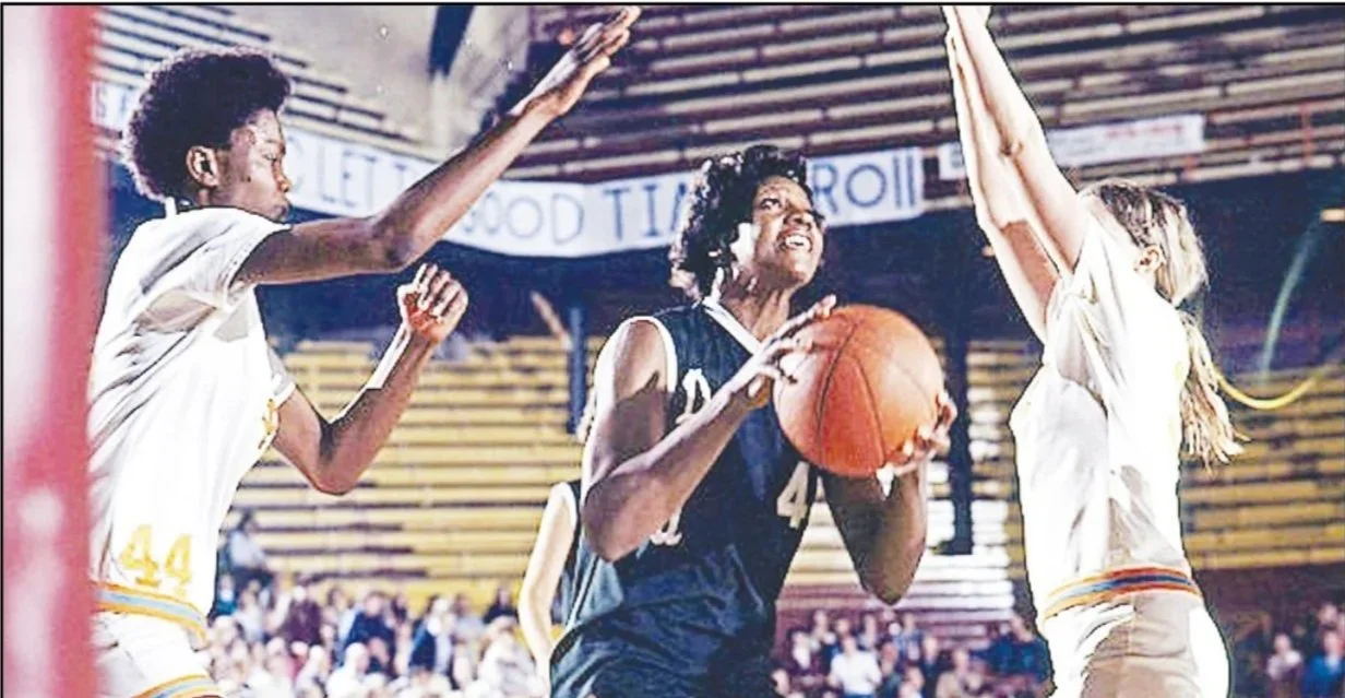 Three female basketball players on the court, one in black jersey holding a basketball and attempting a shot, with two players in white jerseys defending with their arms raised, in an indoor gymnasium.