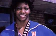 A young woman smiling outdoors, wearing a blue jacket and a black and white striped shirt under a blue hoodie.