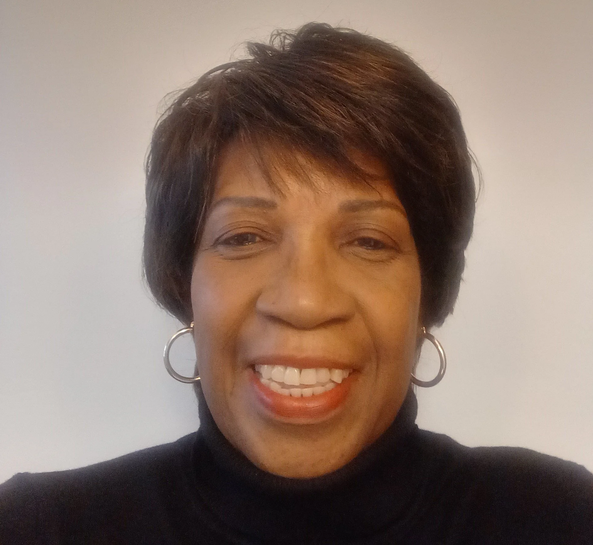 Close-up of a smiling African American woman with short dark hair, wearing hoop earrings and a black turtleneck, against a plain background.