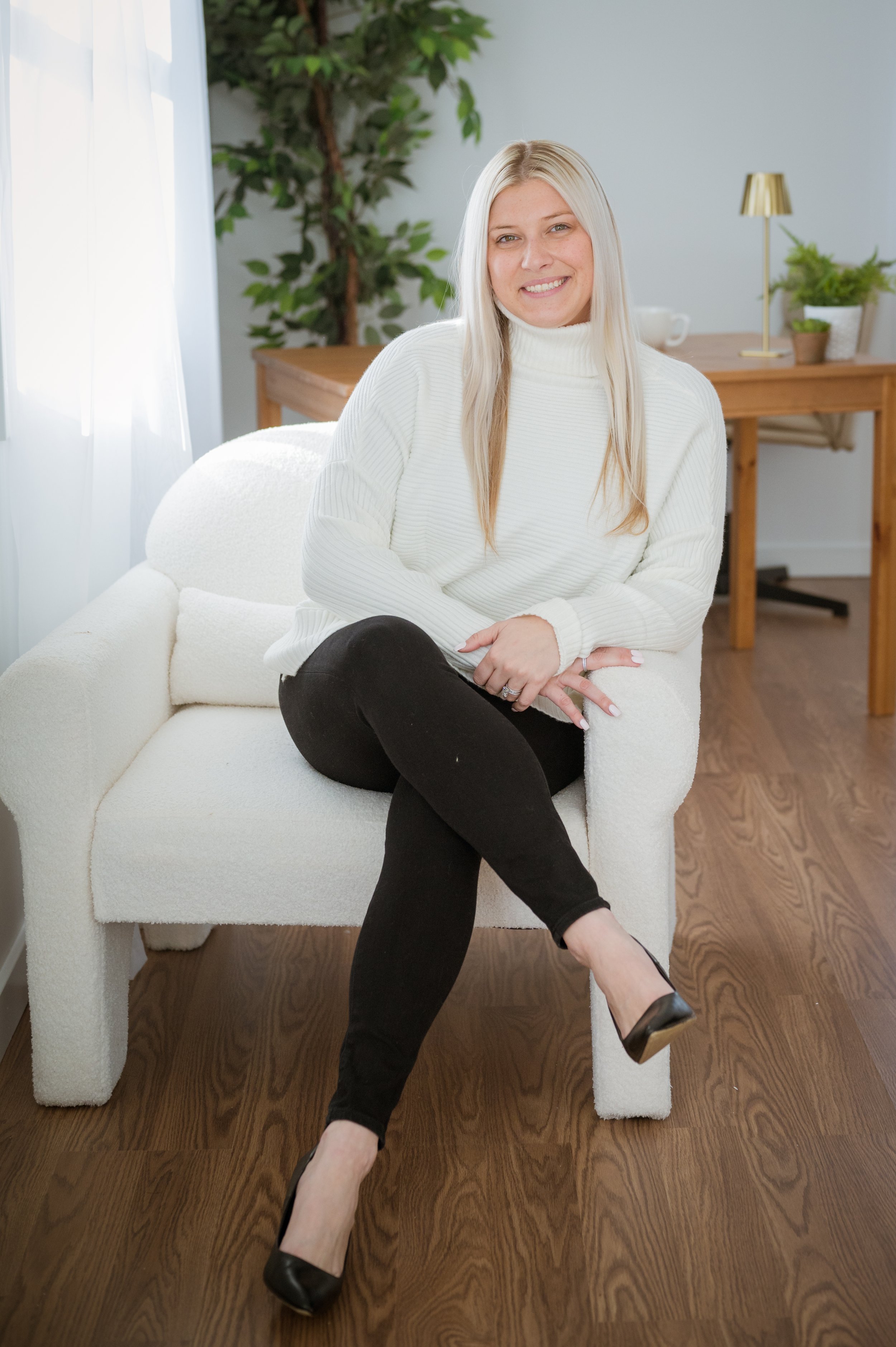 A woman with long blonde hair sitting on a white cushioned armchair in a room with wooden flooring, a wooden table, a potted plant, and a gold-colored lamp in the background. She is wearing a white turtleneck sweater, black pants, and black high heels, and she is smiling at the camera.