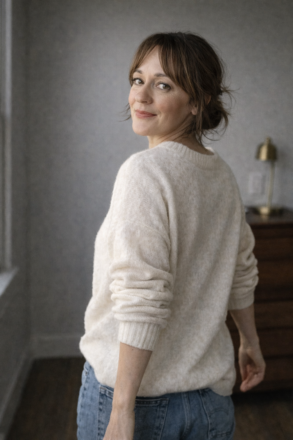 Woman with short brown hair wearing a cream sweater and blue jeans, standing indoors with a grey wall and a brass-colored lamp in the background.