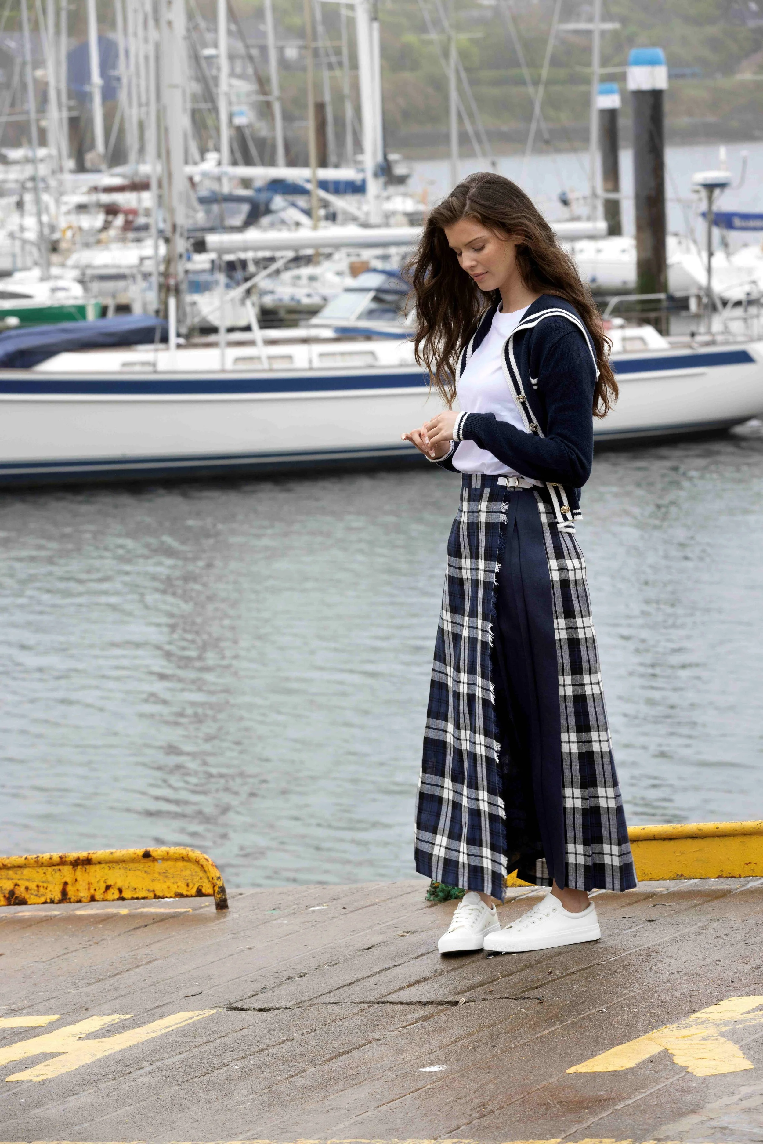 A young woman with long wavy brown hair standing on a dock near the water, looking at her phone. She is wearing a white t-shirt, a dark blue jacket, cream-colored sneakers, and a long plaid skirt. In the background, there are boats and sailboats docked at a marina.